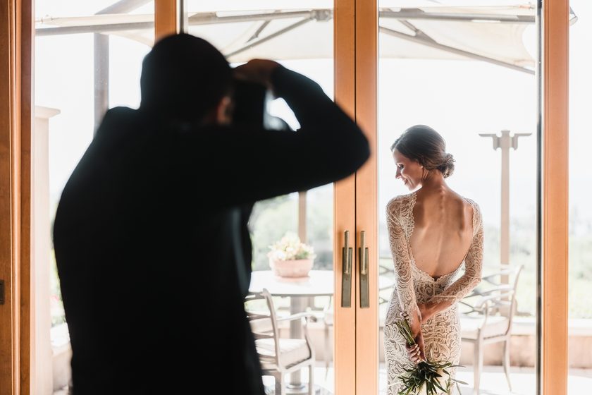 A photographer silhouetted in a doorway, photographing a bride by the window.