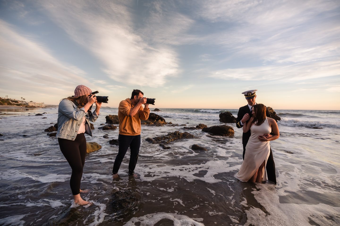 Three photographers standing in the surf at sunset, shooting a couple on the rocks.