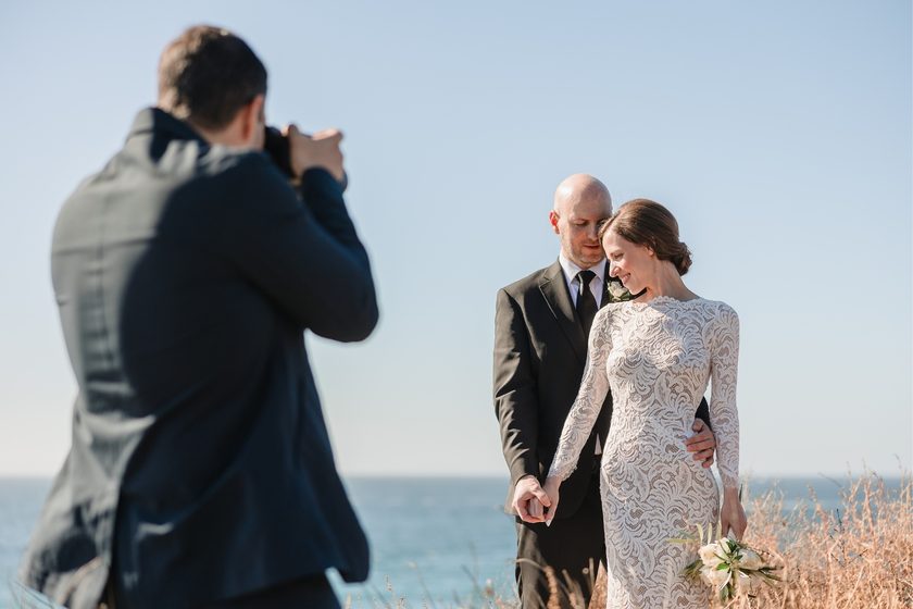 A photographer framing a bride and groom on a coastal clifftop.